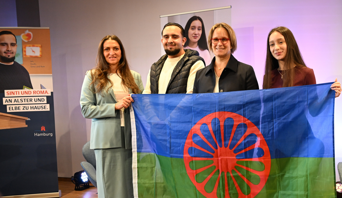 Sinti und Roma zeigen zusammen mit einer Politikerin bei einer Veranstaltung eine Flagge der Roma. Sie setzen sich dafür ein, dass Sinti und Roma nicht ausgegrenzt werden. 