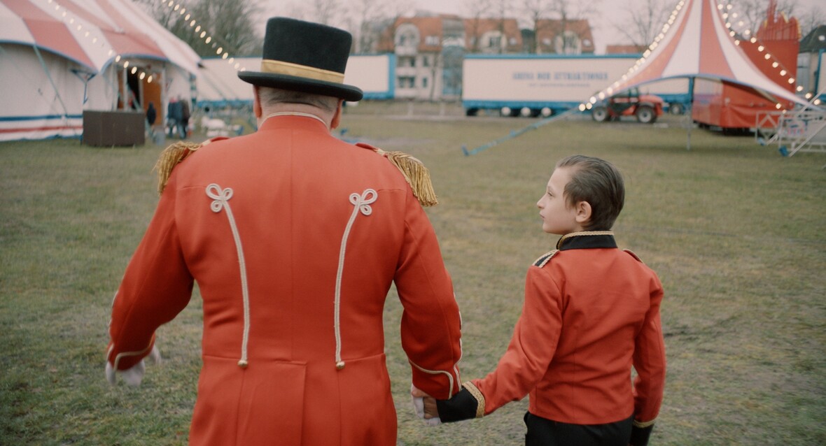 Santino und sein Opa gehen Hand in Hand über den Platz. Sie tragen Zirkusuniform