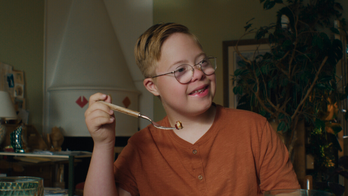 Lars sitzt am Küchentisch und hält eine Gabel mit Essen in der rechten Hand. Er lächelt. 