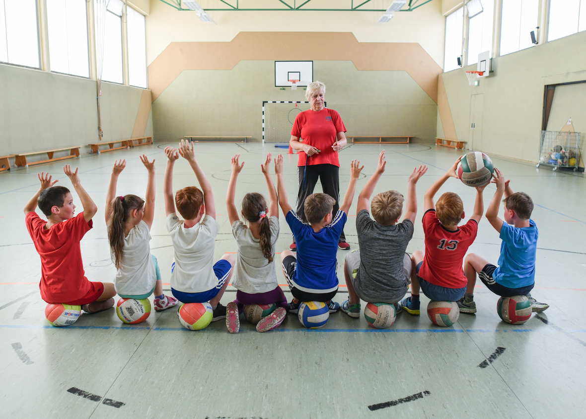 Kinder treiben Sport in einer Grundschule in Strausberg. Man sieht sie von hinten, während die auf Medizinbällen sitzen und zu ihrer Lehrerin schauen.