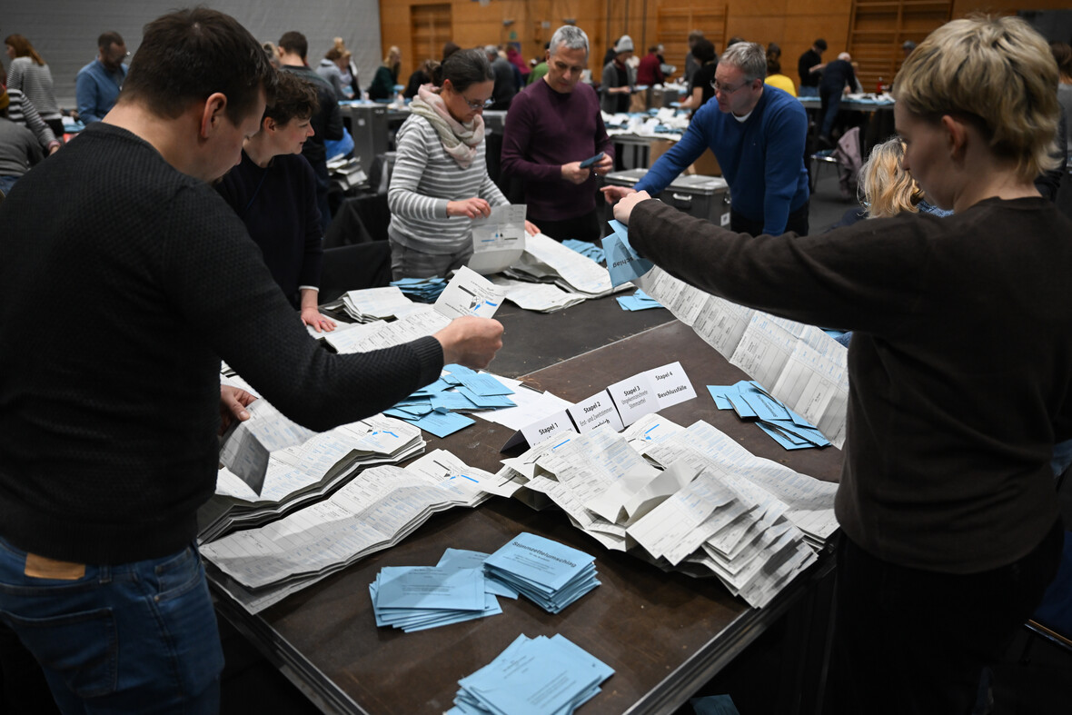 Wahlhelfer/-innen zählen Stimmen aus. Sie stehen an einem Tisch und schauen sich die Stimmzettel genau an. 